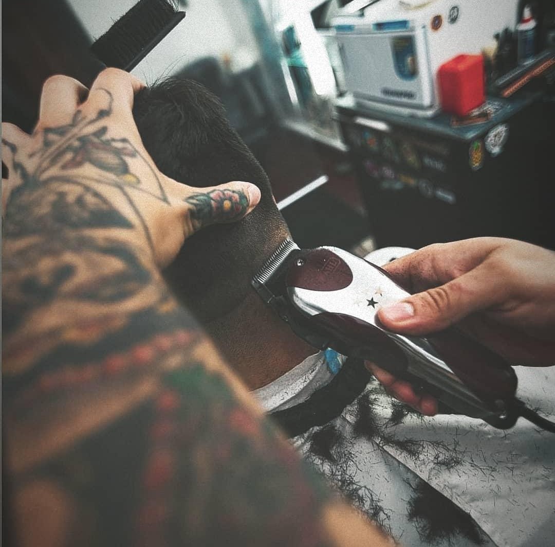 Action shot of a professional barber performing a precision haircut in a high-end, moody studio setting. The lighting is warm and directional, highlighting the detail of the tools and the focus of the artist. The background features dark textures and premium studio equipment, creating an atmosphere of masculine luxury and expert craftsmanship.